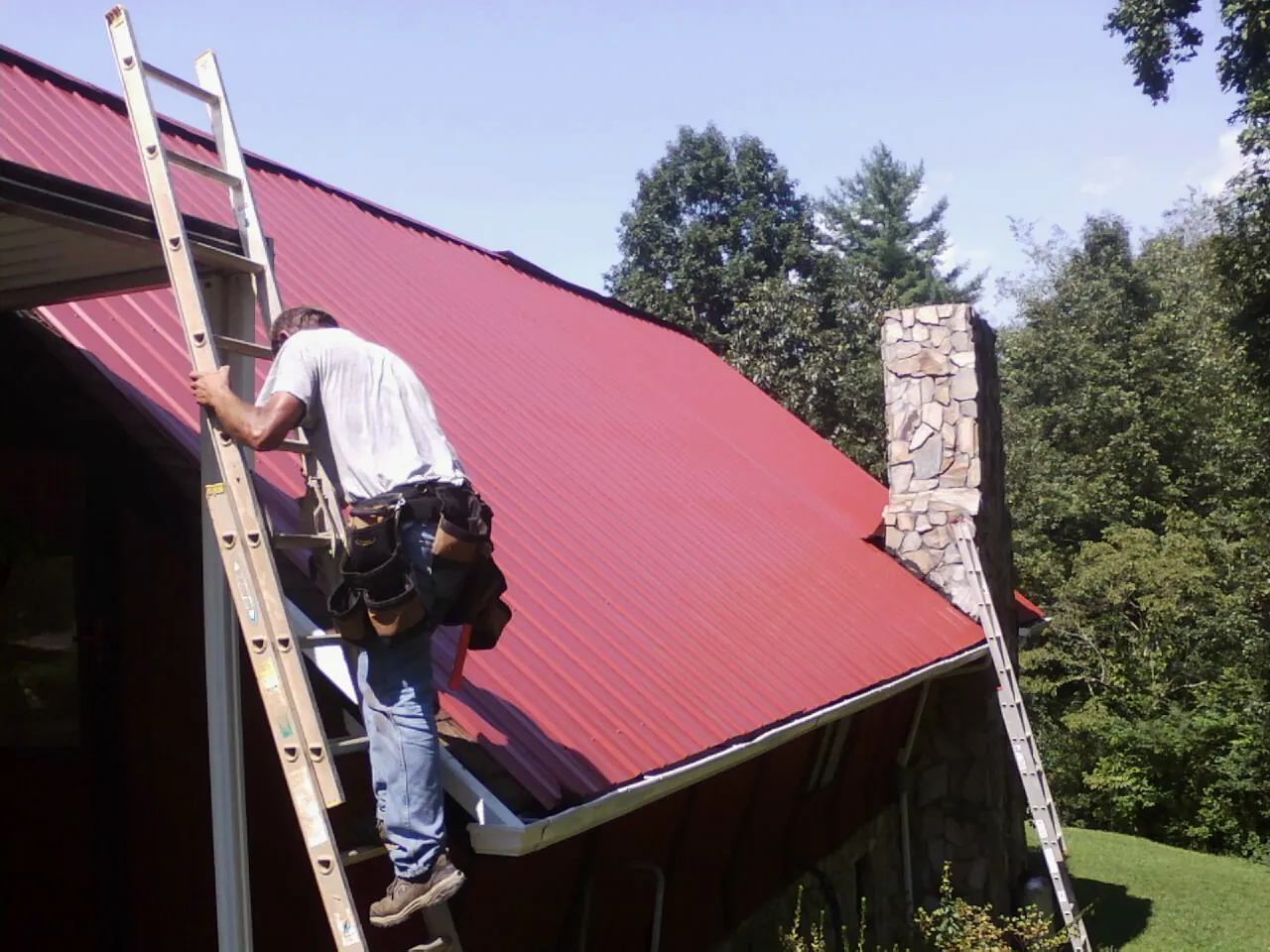 Roofer on a ladder beside a red metal roof and stone chimney — Soffit & Fascia Repair by Roofing Brothers in Holland